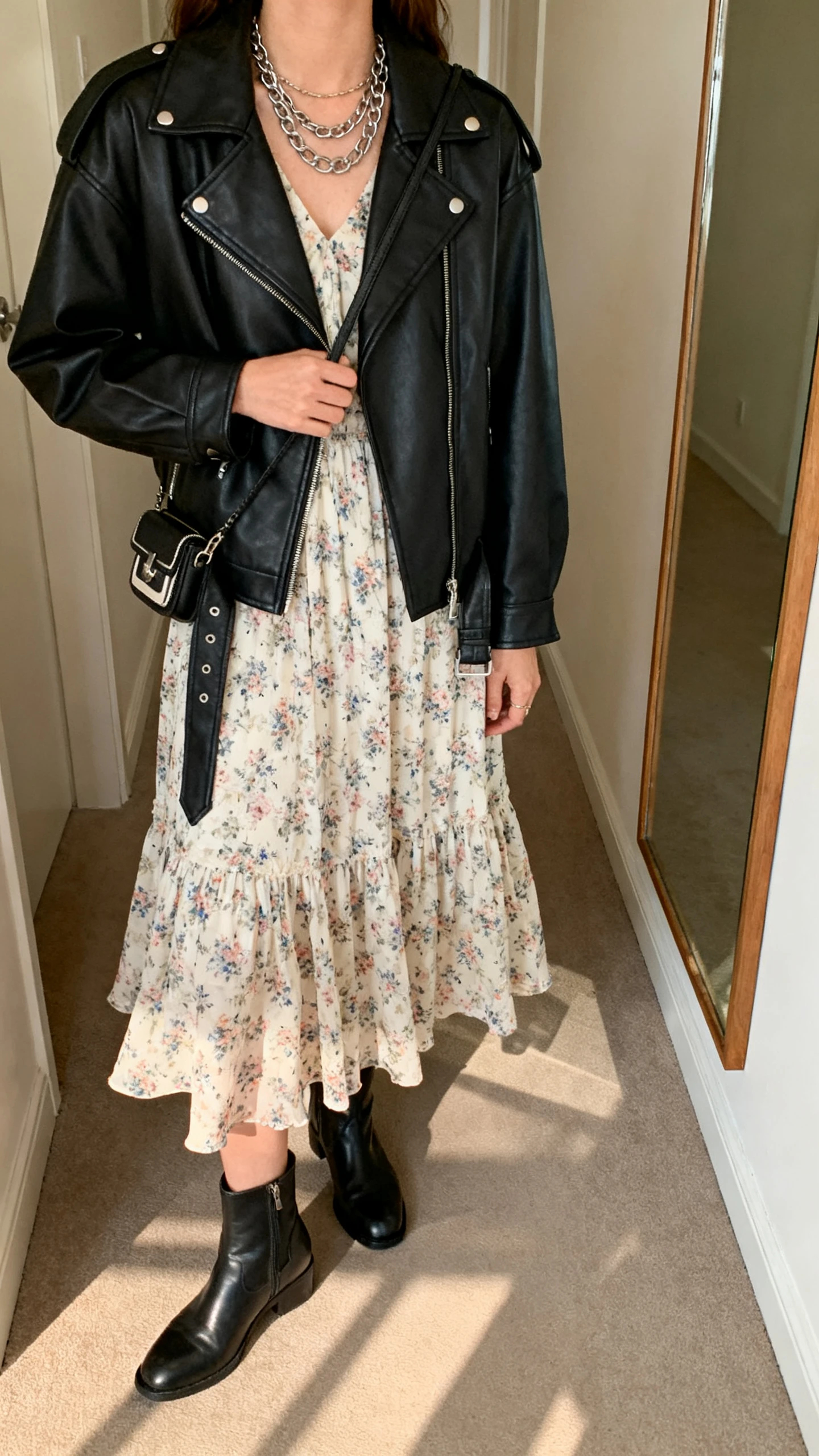 iPhone back/side view of a woman in an edgy black leather jacket over a floaty floral midi dress, ankle boots, layered chains, and a mini crossbody, face not visible, hallway mirror, indoor warm light, iPhone photo quality.