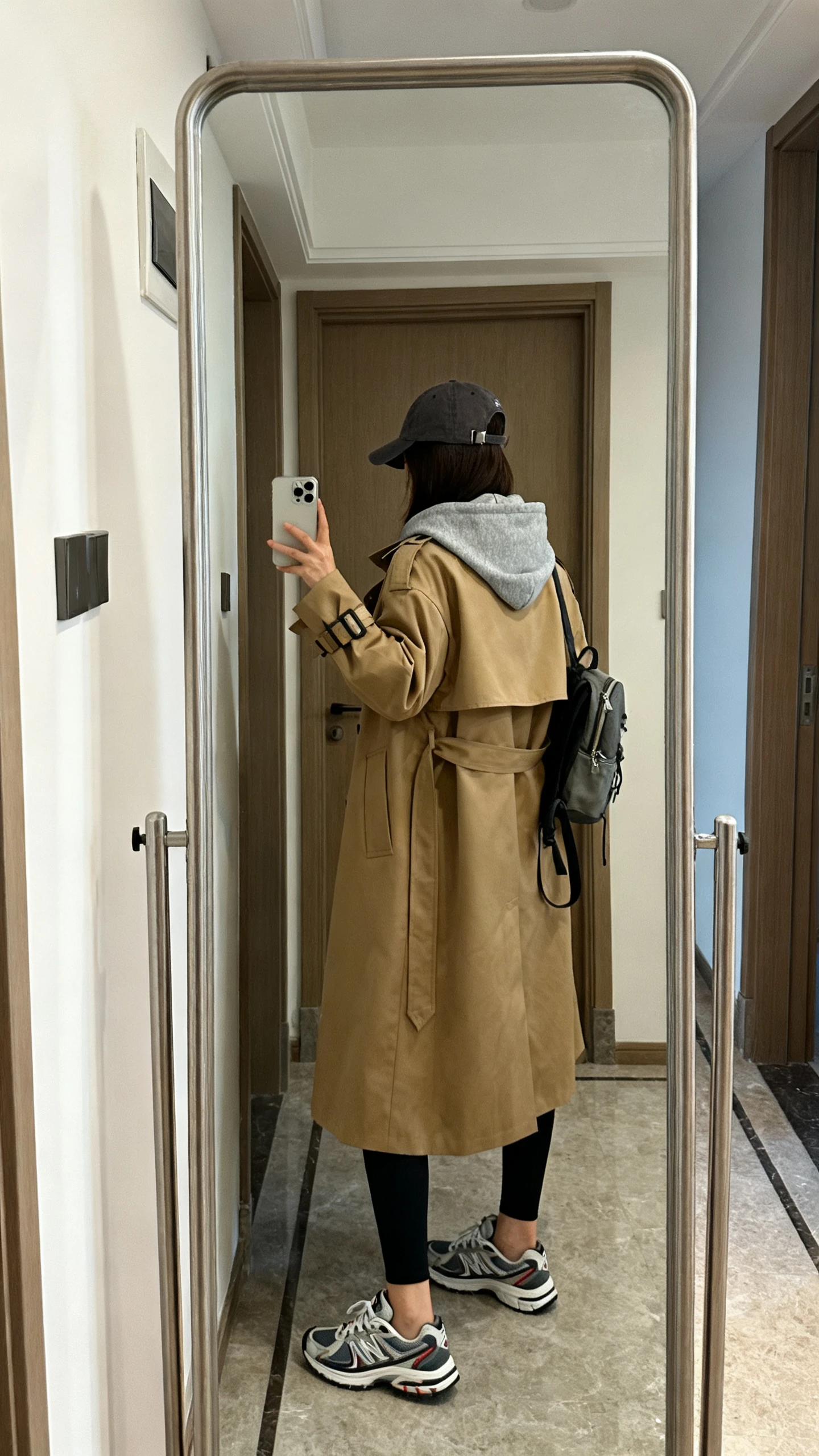 iPhone back-view mirror selfie of a woman wearing a gray hoodie layered under a classic tan trench coat, black leggings, baseball cap, and retro running sneakers, small backpack, face not visible, entryway mirror, natural daylight, iPhone photo quality.