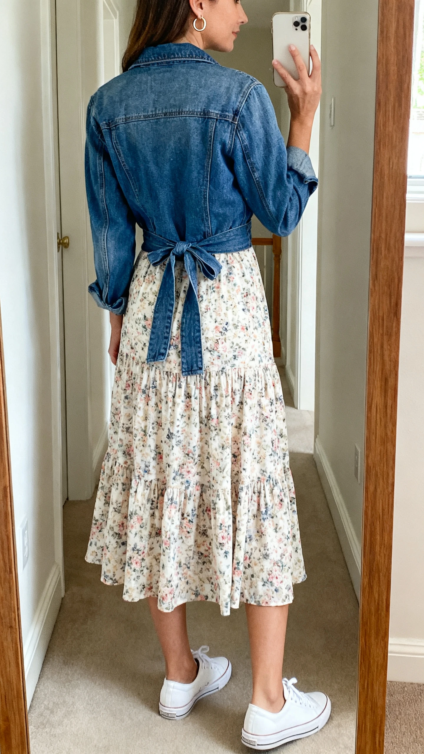 iPhone back-view selfie of a woman wearing a breezy floral midi dress with white low-top sneakers and a denim jacket tied at the waist, small hoop earrings, face not visible, hallway full-length mirror, soft natural daylight, iPhone photo quality.