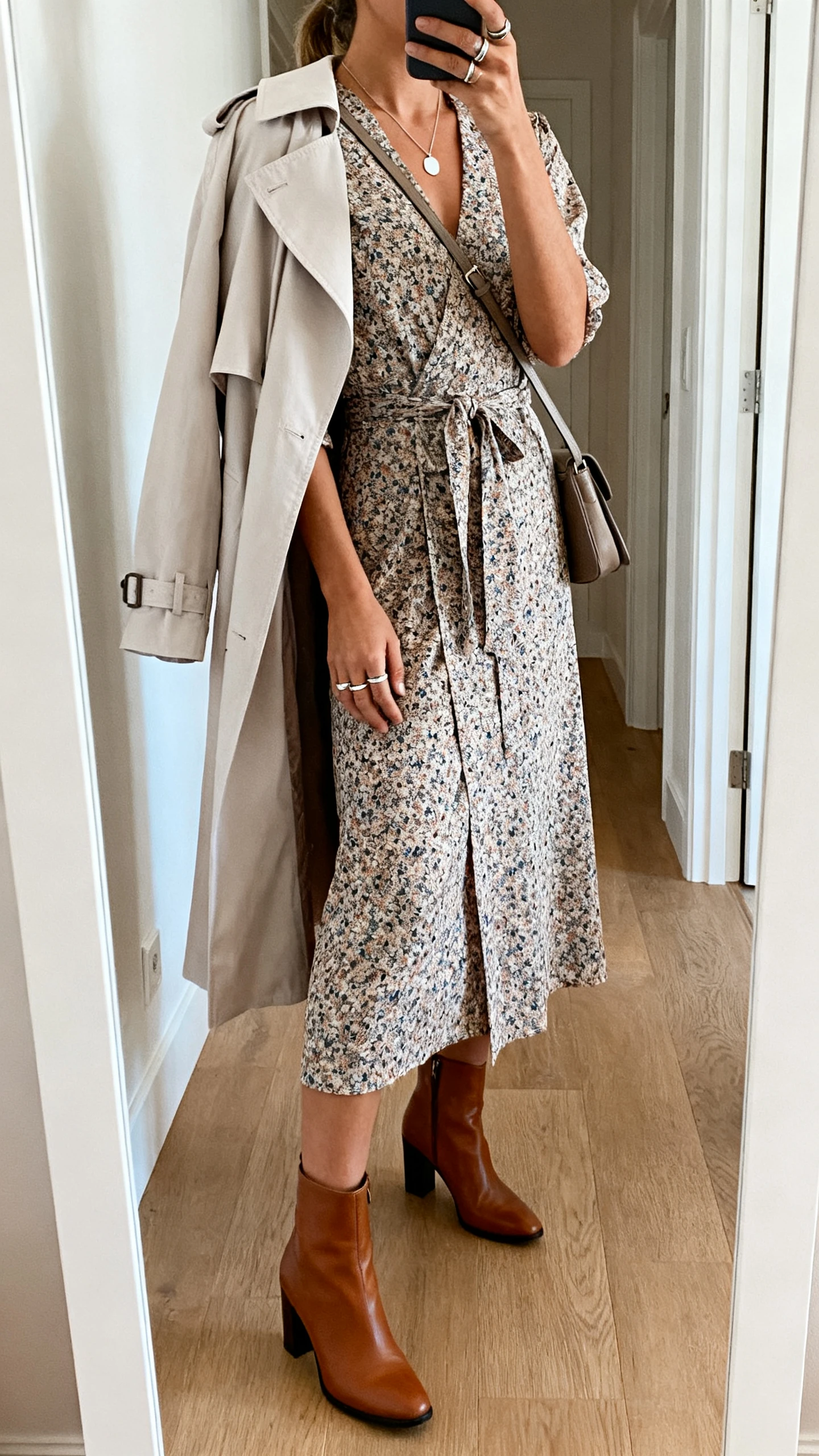 iPhone back-view selfie of a woman wearing a printed midi wrap dress with leather mid-heel ankle boots, a light trench coat draped over shoulders, minimal pendant necklace, stacked rings, and a crossbody bag; face not visible, hallway full-length mirror, natural daylight, iPhone photo quality.