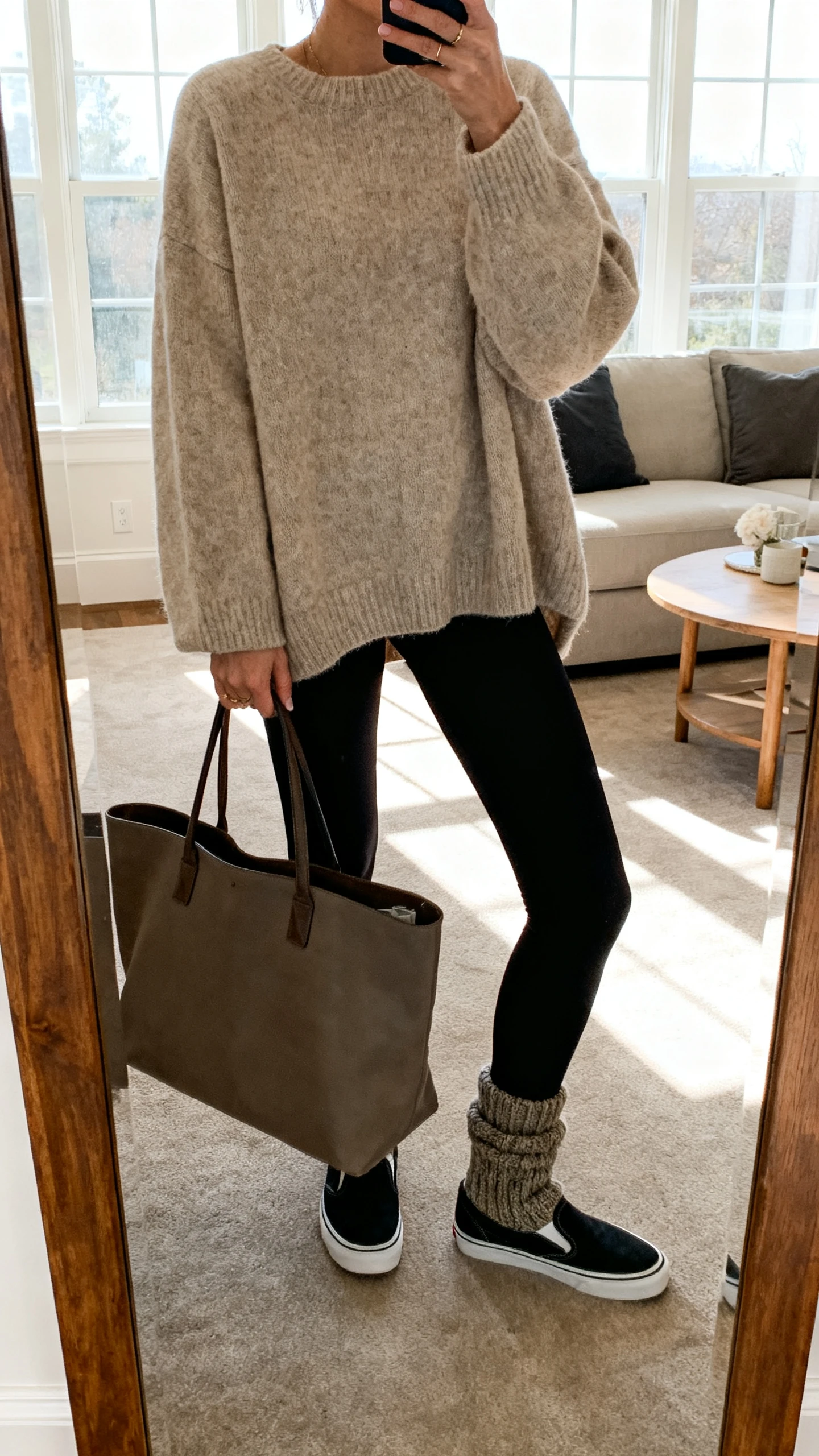iPhone mirror selfie of a woman in a cozy oversized oatmeal sweater with black leggings, chunky socks, slip-on sneakers, and a big tote, face not visible, cropped at neck, living room mirror, morning window light, iPhone photo quality.