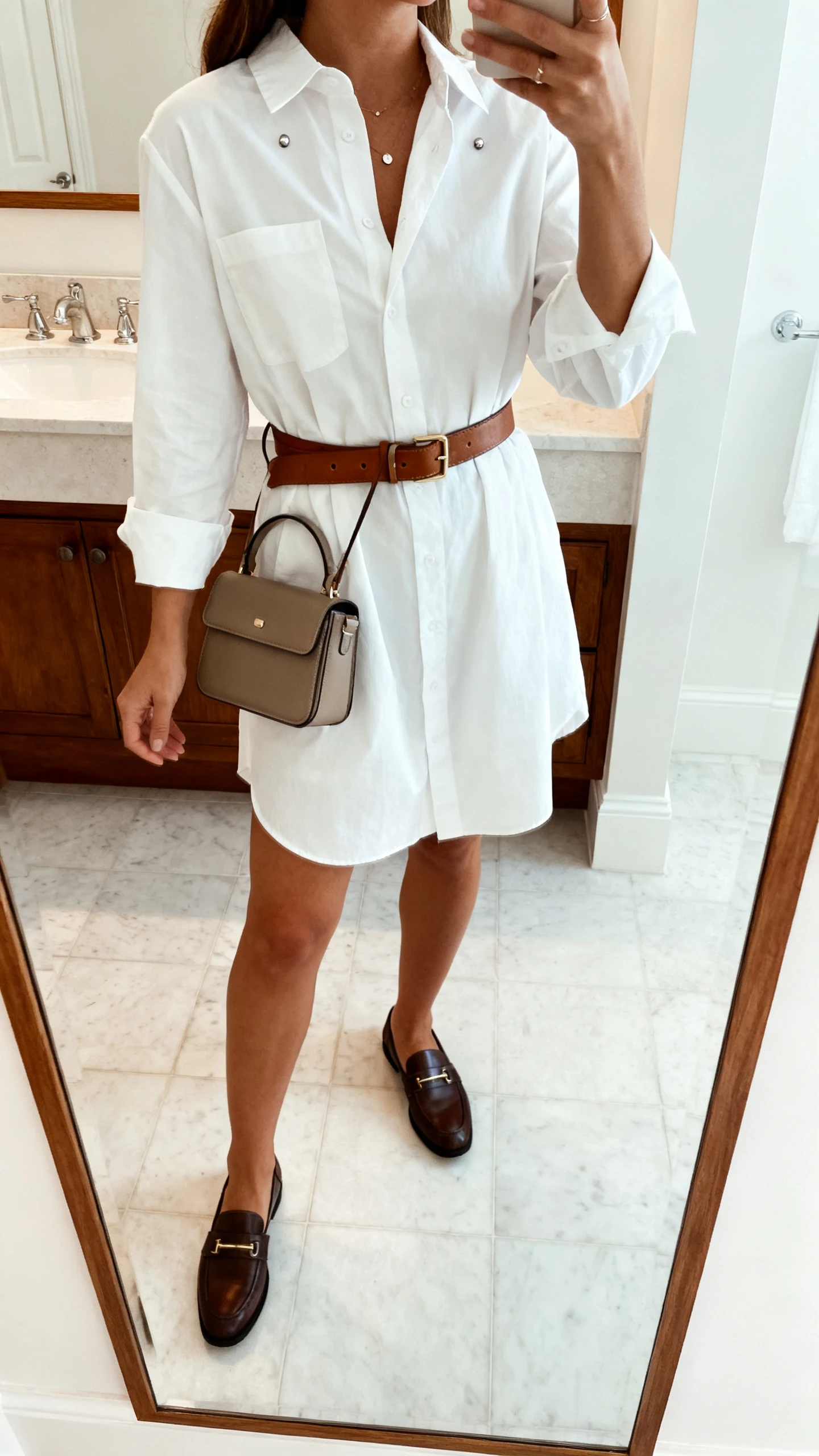 iPhone mirror selfie of a woman in a crisp white shirt dress cinched with a leather belt, loafers, structured mini bag, and simple studs, face not visible, cropped at shoulders, bathroom mirror, bright indoor light, iPhone photo quality.