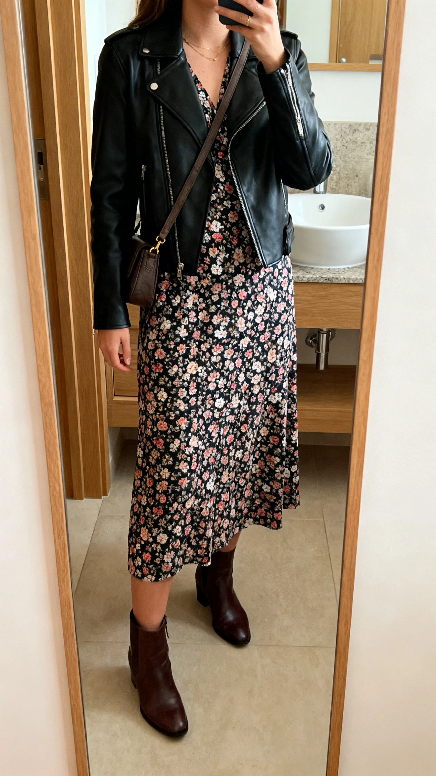 iPhone mirror selfie of a woman in a floral midi dress under a black leather jacket, ankle boots, and a crossbody bag, face not visible, cropped at shoulders, bathroom mirror, warm indoor lighting, iPhone photo quality.