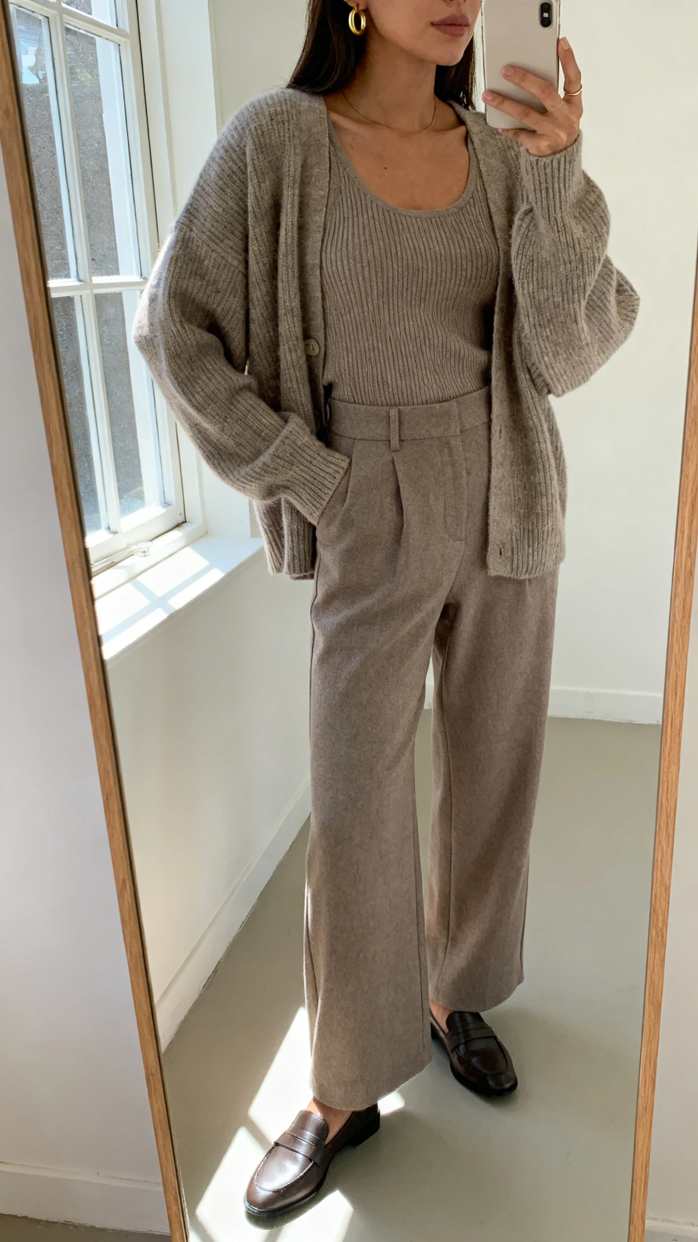 iPhone mirror selfie of a woman in a monochrome look: tonal knit sweater and matching knit tank, wide-leg trousers in the same shade, sleek loafers, and delicate gold hoops, face not visible, cropped at shoulders, full-length mirror by a window, natural window light, iPhone photo quality.