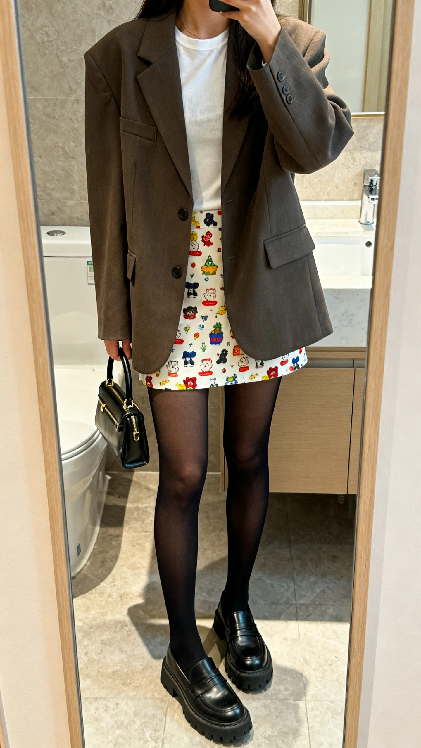 iPhone mirror selfie of a woman in a playful mini skirt with opaque black tights, boxy oversized blazer over a fitted tee, chunky loafers, and a small top-handle bag, face not visible, bathroom mirror, soft indoor lighting, iPhone photo quality.