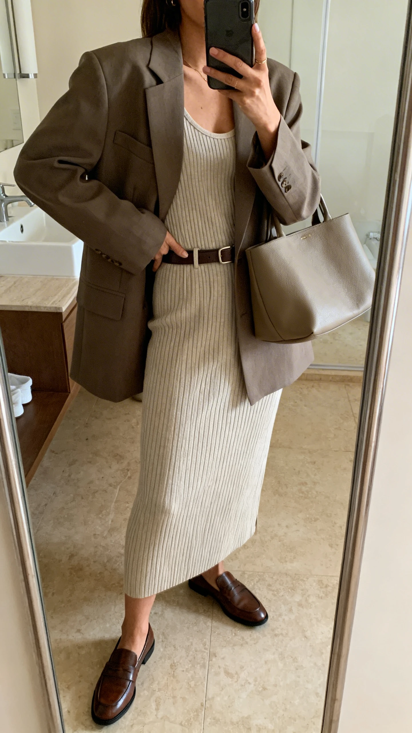 iPhone mirror selfie of a woman in a tailored oversized blazer layered over a ribbed knit tank and matching ribbed knit skirt, with leather loafers, a slim belt, and a roomy tote; face not visible, side angle, bathroom mirror, soft indoor lighting, iPhone photo quality.