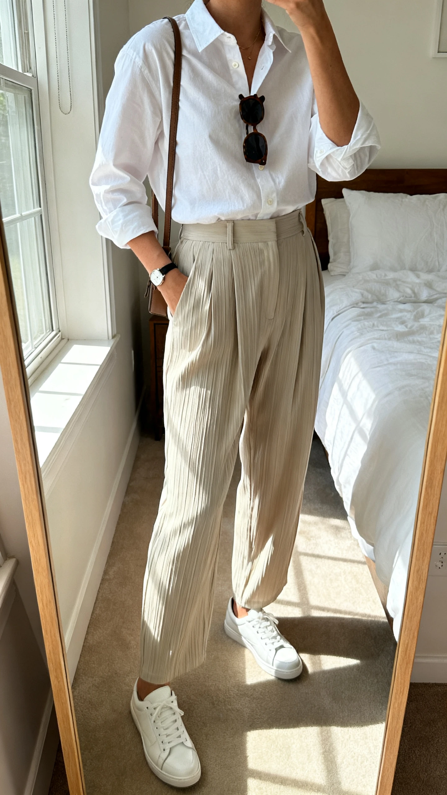 iPhone mirror selfie of a woman in a white cotton button-down tucked into high-rise relaxed pleated trousers, clean white leather sneakers, understated crossbody, classic sunglasses hooked at neckline, and a slim watch; face not visible, cropped at shoulders, bedroom mirror, morning window light, iPhone photo quality.