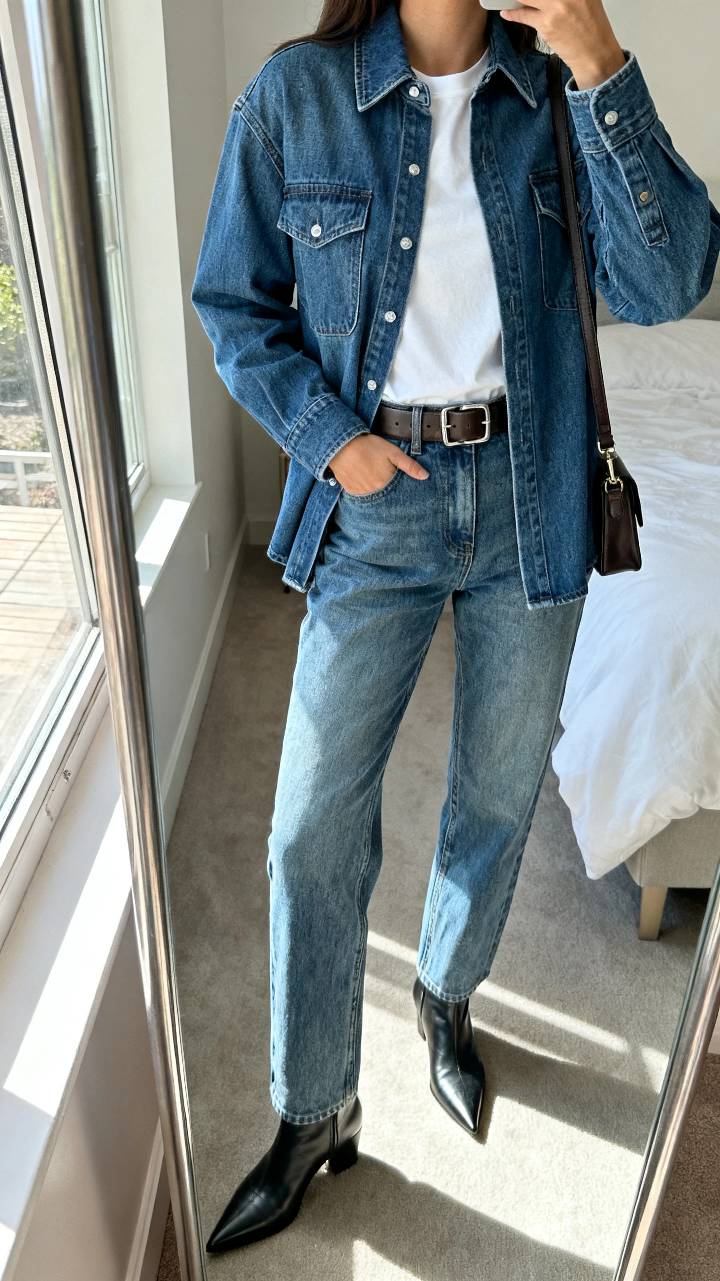 iPhone mirror selfie of a woman in denim-on-denim: medium-wash relaxed jeans with a matching structured denim shirt jacket, white tee underneath, sleek leather belt, pointed ankle boots, and a minimal crossbody, face not visible, cropped at shoulders, bedroom mirror, natural window light, iPhone photo quality.