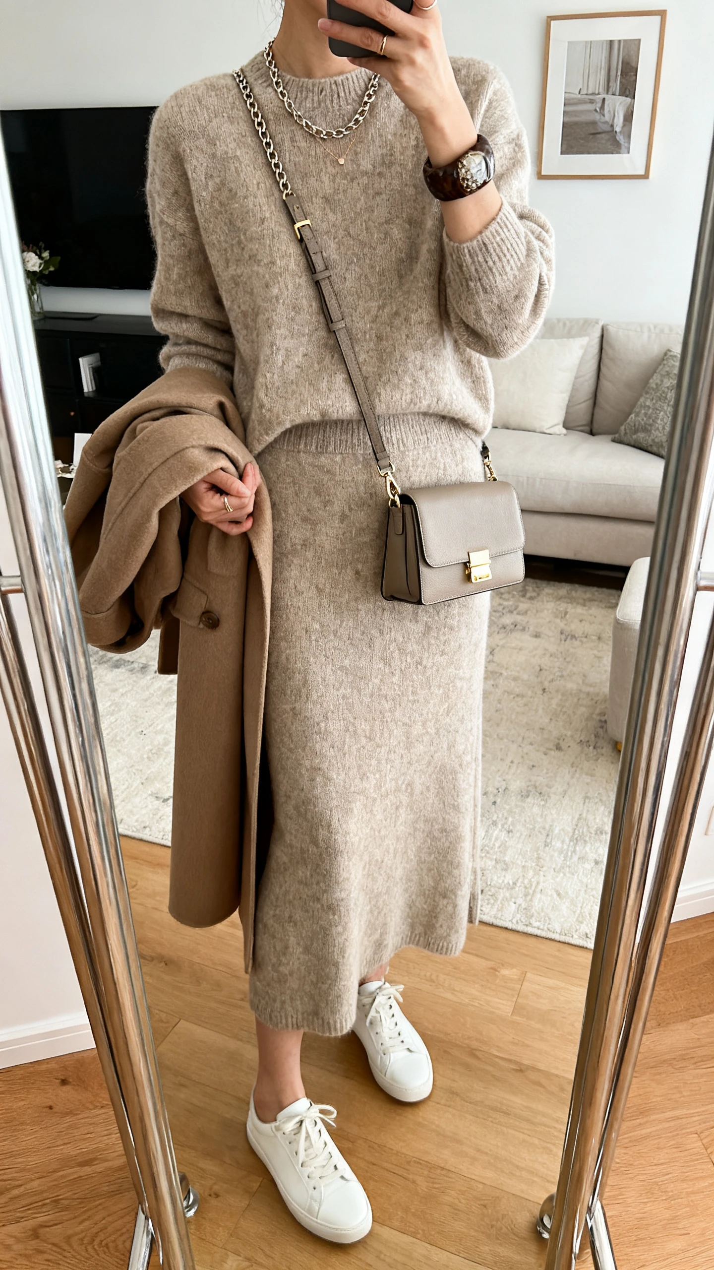 iPhone mirror selfie of a woman wearing a coordinating knit sweater and midi skirt set with minimal white sneakers, a structured crossbody, delicate chain necklace, chunky cuff, and an optional longline coat draped over arm; face not visible, cropped at neck, living room full-length mirror, indoor natural light, iPhone photo quality.