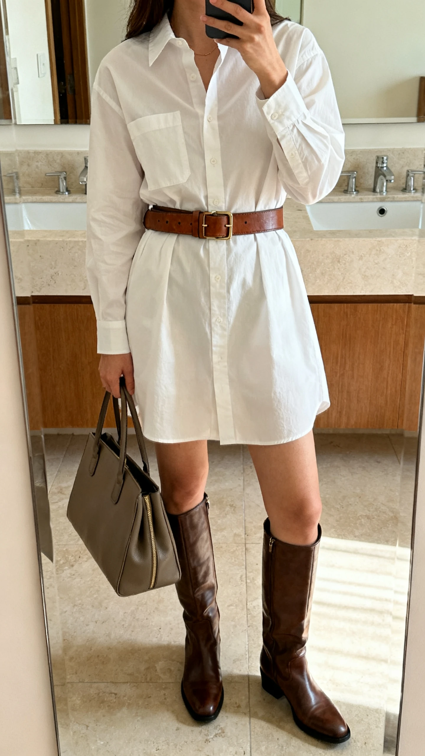 iPhone mirror selfie of a woman wearing a crisp shirt dress cinched with a leather belt, knee-high boots, and a structured tote, face not visible, cropped at shoulders, bathroom mirror, bright indoor lighting, iPhone photo quality.