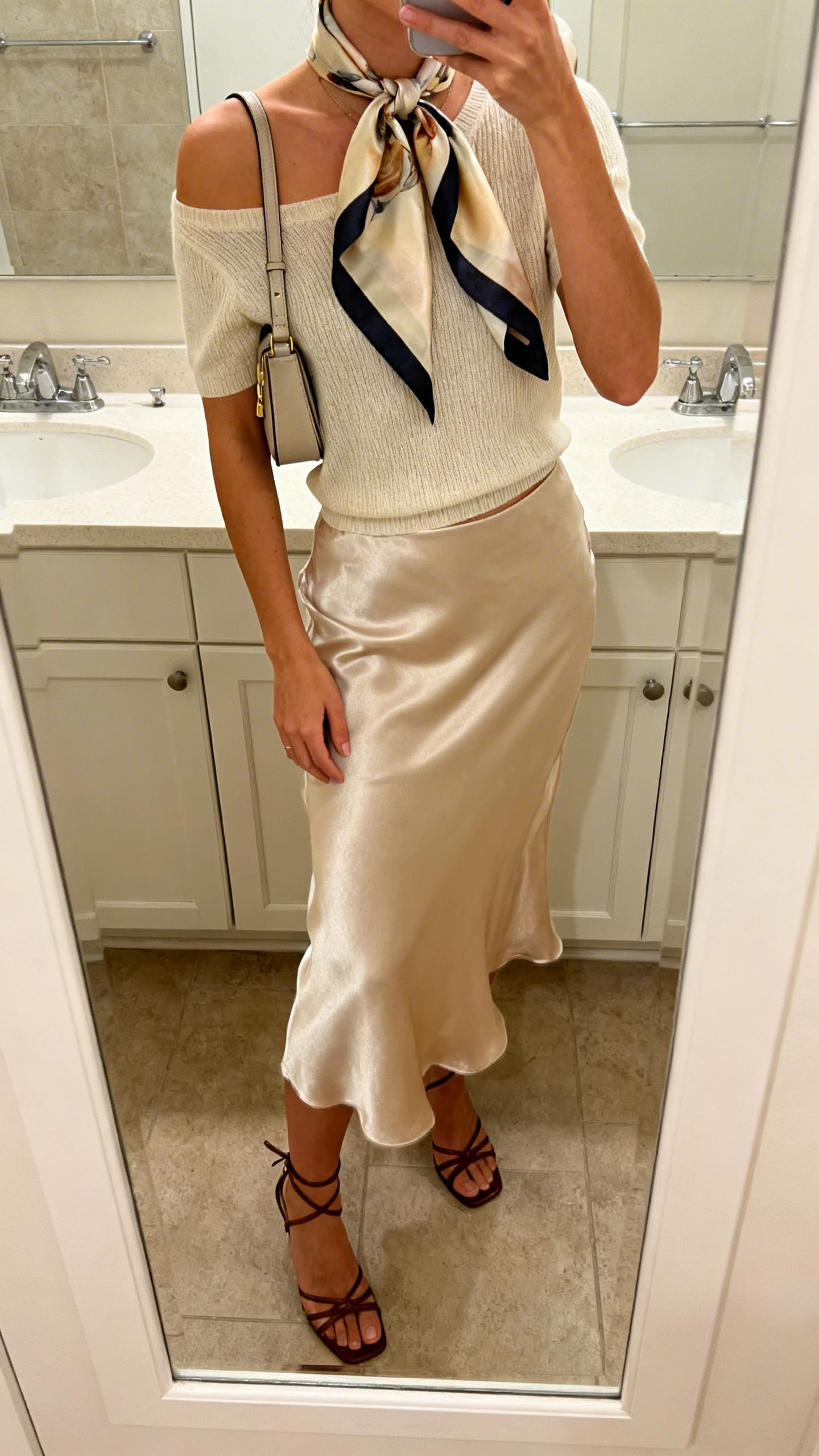 iPhone mirror selfie of a woman wearing a silk scarf tied at the neck, satin slip skirt, minimal knit top, strappy heels, and a small shoulder bag, face not visible, cropped at shoulders, bathroom mirror, warm indoor light, iPhone photo quality.