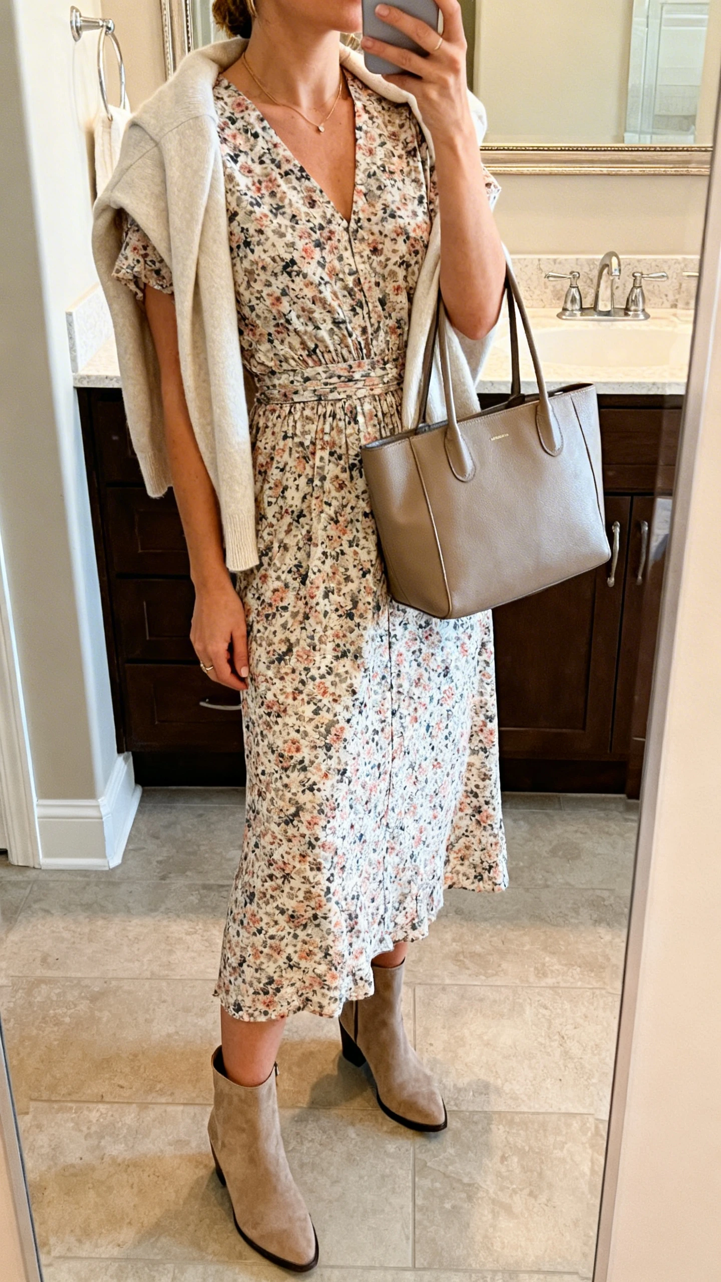 iPhone side-angle mirror selfie of a woman wearing a floral midi dress with a defined waist, ankle boots, a light cardigan draped over shoulders, and a structured tote, face not visible, bathroom mirror with soft indoor lighting, iPhone photo quality.