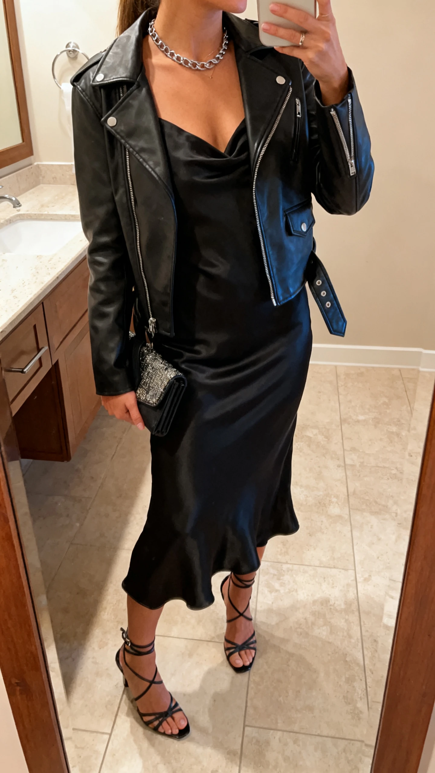 iPhone side-angle mirror selfie of a woman wearing a silky black slip dress layered with a black leather moto jacket, strappy heels, delicate chain necklace, and a clutch, face not visible, bathroom mirror, warm indoor lighting, iPhone photo quality.